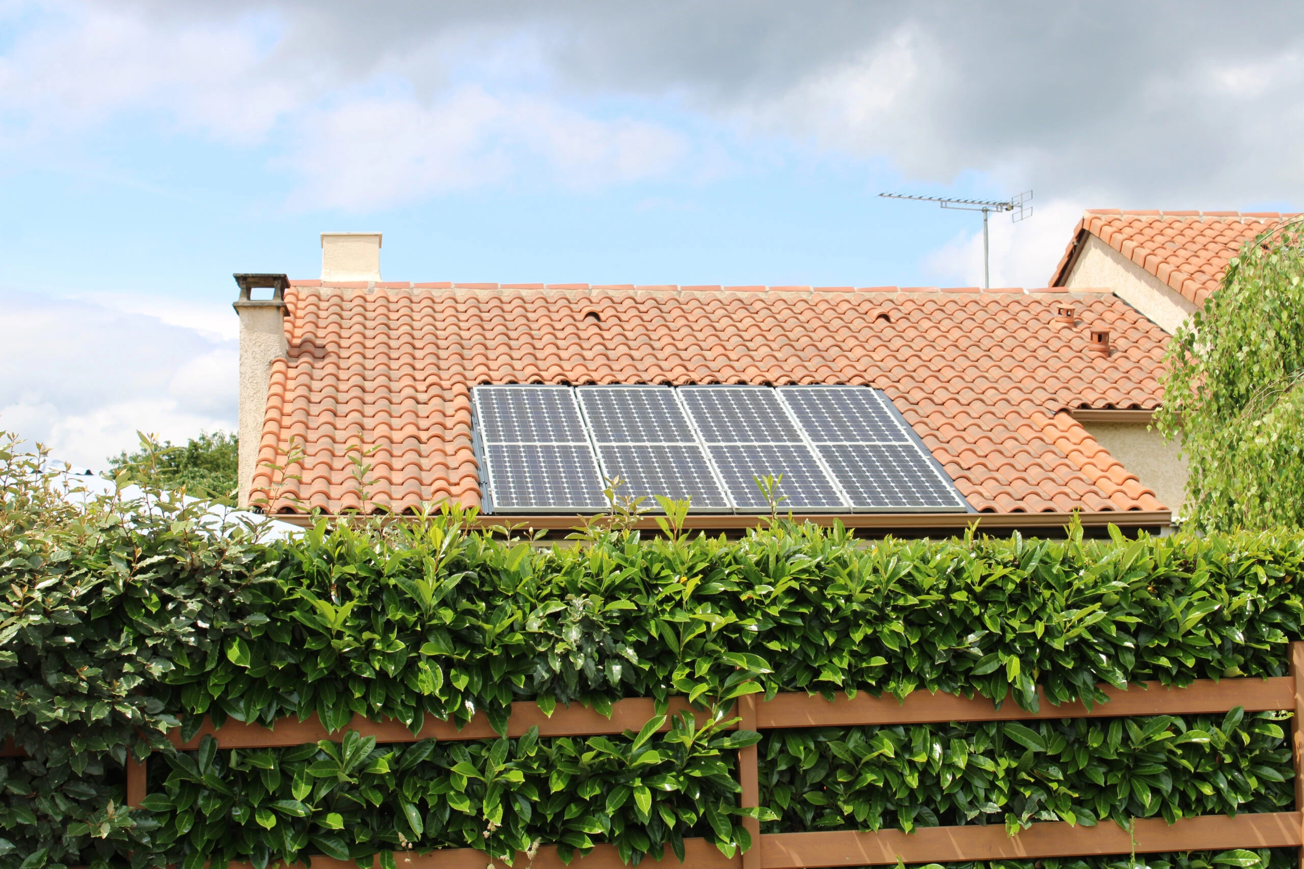 Maison au toit de tuiles équipée de panneaux solaires derrière une haie